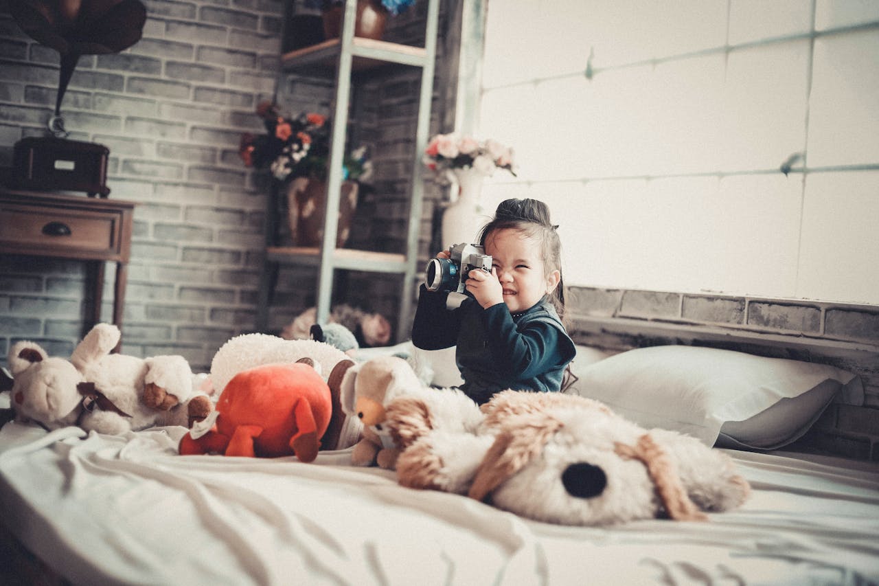 Smiling child with a camera surrounded by stuffed toys in a cozy bedroom setting.